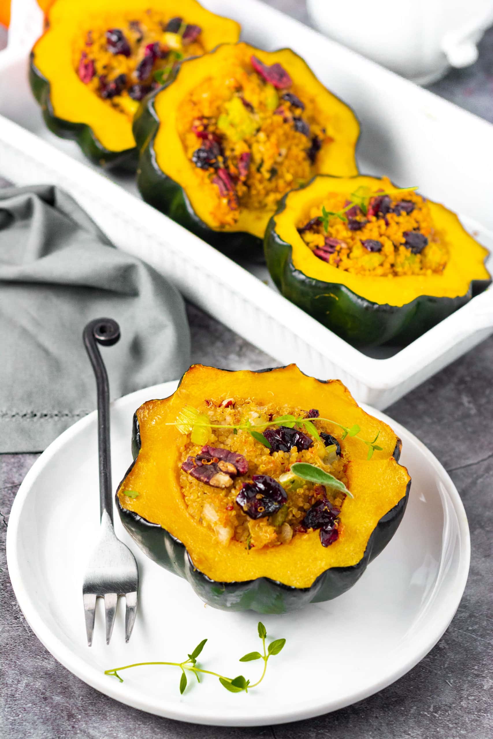 Vegan Stuffed acorn squash on a white plate stuffed with quinoa, cranberries, pecans, and sausage with a black fork with a white casserole dish in the background