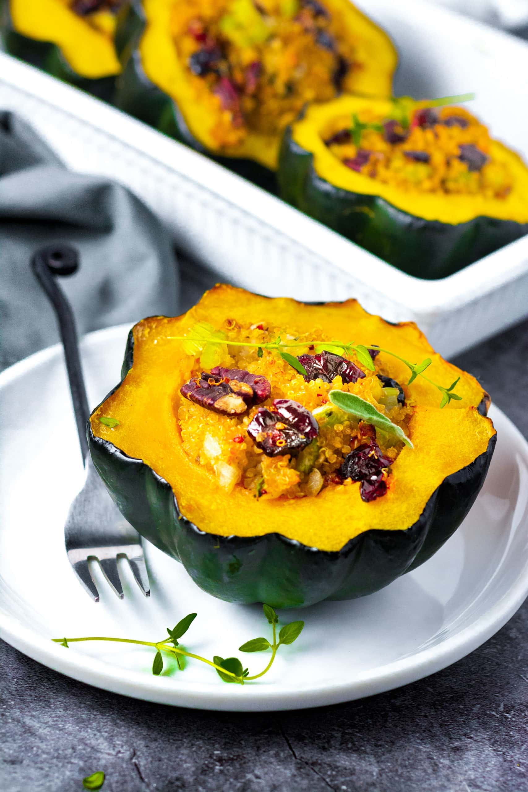 Vegan Stuffed acorn squash on a white plate stuffed with quinoa, cranberries, pecans, and sausage with a black fork with a white casserole dish in the background