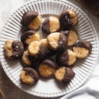 Estos deliciosos Bocaditos Helados de Crema de Almendra Plátano y Chocolate, son un snack perfecto. Son unos caprichitos saludables ideales para los días de verano y, ya que son del tamaño de un bocado, podemos considerarlos bajos en calorías (si logras no comerte más de 1 o 2 a la vez). Foto desde encima donde se ve un palto con todos los bocaditos en el centro, abajo a la izquierda sal, arriba a la derecha chocolate derretido en una taza.