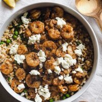 Ensalada Tibia de Gambas y Cebada: súper deliciosa, nutritiva y fácil de preparar. Tiene toques de limón, ajo y pimentón, con guisantes y feta, que se deshace al mezclarlo con las gambas calientes para redondear el plato. En esta foto cenital se ve la ensalada servida en un bol con gajos de limón arriba a la izquierda, cubiertos de madera para servir la ensalada arriba a la derecha y un pequeño bol con guindillas abajo a la derecha.