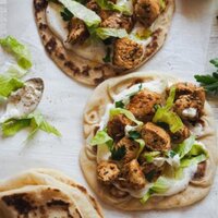 This Mediterranean Spiced Chicken Wraps with Cream Cheese Sauce and Flatbread is so juicy! Chicken marinated with Mediterranean herbs and a spicy and somky touch. Then grilled and served with a delicious cream cheese sauce and Turkish flatbread. It makes a great meal served like a wrap or pita. This is an overhead shot of two flatbreads layered with cream cheese sauce, some spiced chicken, the lettuce and parsley. There is a folded flatbread on the bottom left corner and some lettuce on the bottom right.