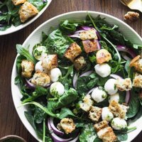 Esta ensalada de espinacas con vinagreta de alcaparras combina todos los ingredientes clave de una buena ensalada: verde del bueno con espinacas y rúcula, crujiente con picatostes, color con el contraste blanco de la mozzarella y el morado de la cebolla, todo ligado con una deliciosa vinagreta de alcaparras. Si tienes picatostes a mano, en 5 minutos la tienes lista! Foto cenital del plato de ensalada. Este está en el centro y arriba a la izquierda se aprecia un trocito de otro plato con más ensalada. Arriba a la derecha hay una cuchara y tenedor dorados rodeados por picatostes. Abajo a la derecha hay un pequeño bol con aliño extra.