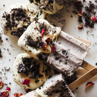 Helado Cremoso de Oreo con Solo 3 Ingredientes, perfecto para combatir el calor y matar antojos de dulce. La cobertura de chocolate blanco y oreo es totalmente opcional. Así que si quieres evitarte un par de pasos e ingredientes, puedes congelar el helado en un recipiente hermético o molde directamente, y es que tal cual ya está buenísimo. Foto con ángulo donde se ven unos cuantos polos de este helado de oreo bañados en chocolate blanco y con oreo troceada por encima.| Receta de Paula's Apron