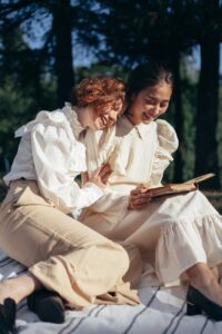 smiling women in old fashioned clothing reading book on picnic blanket