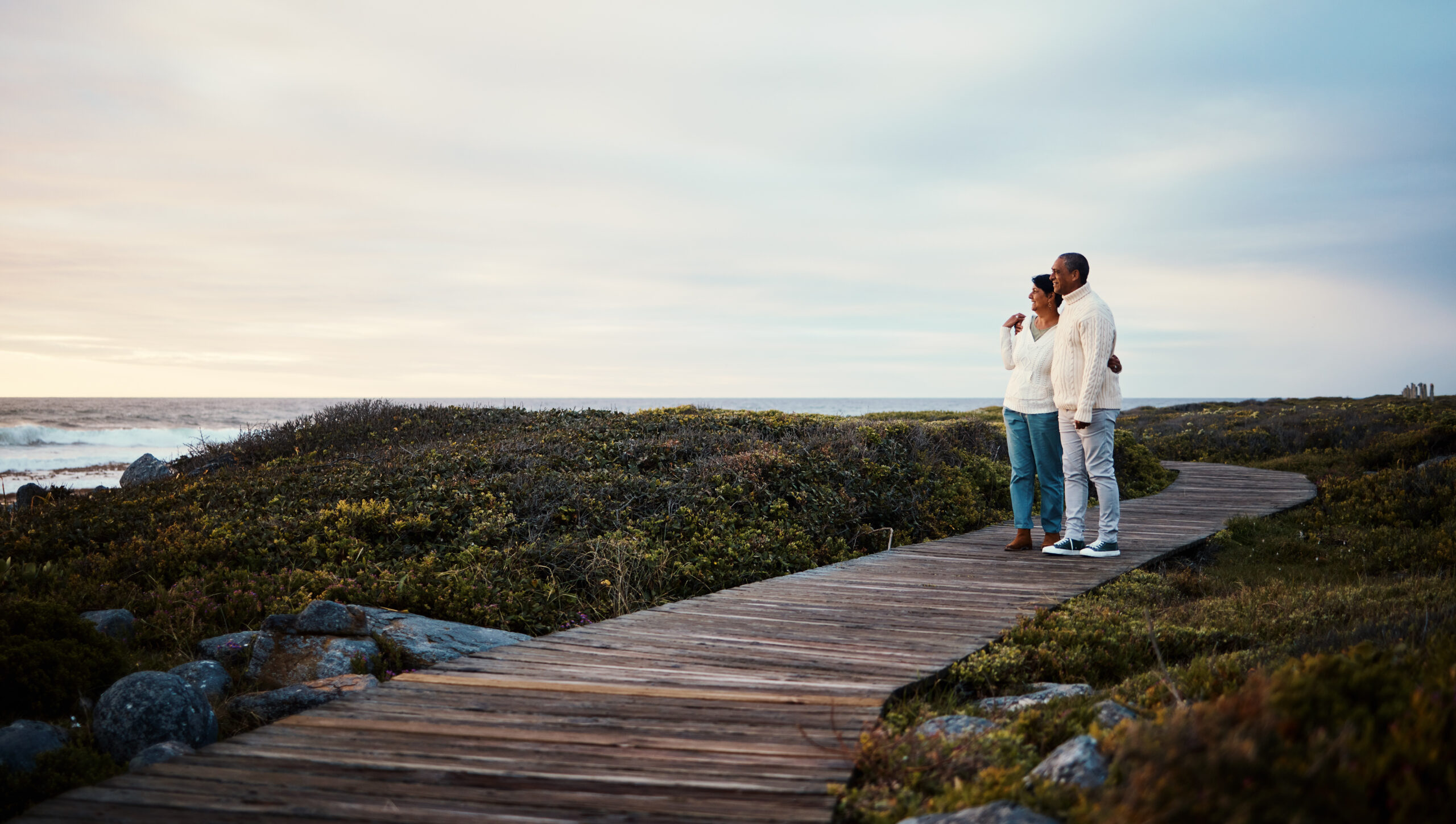 Love, travel and elderly couple hug on a boardwalk at a beach, calm and content against blue sky background. Senior, man with woman on ocean trip, holiday or vacation, happy and enjoying retirement.