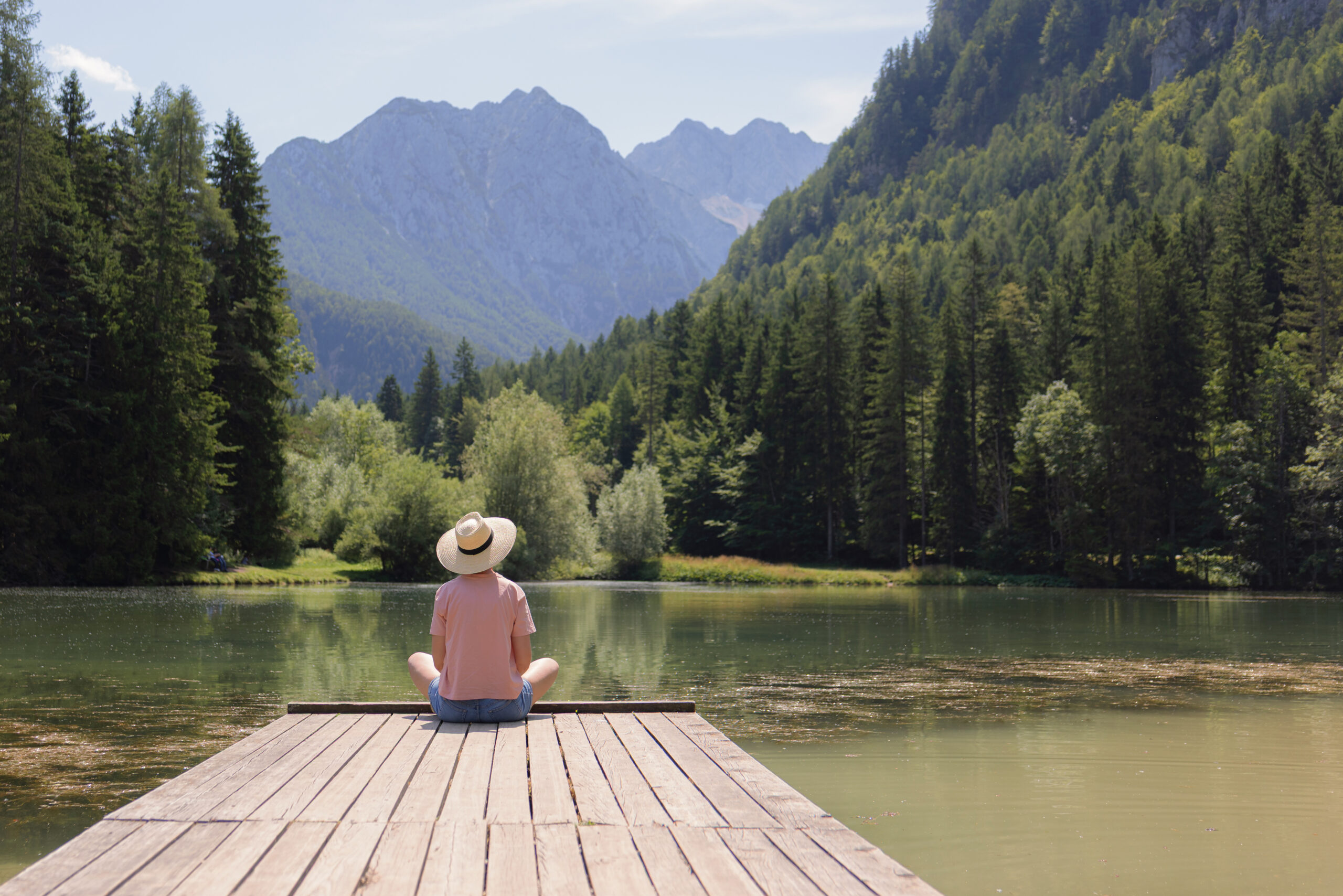 A person sits peacefully on a wooden dock by the lake, surrounded by mountains and dense greenery under a clear blue sky.