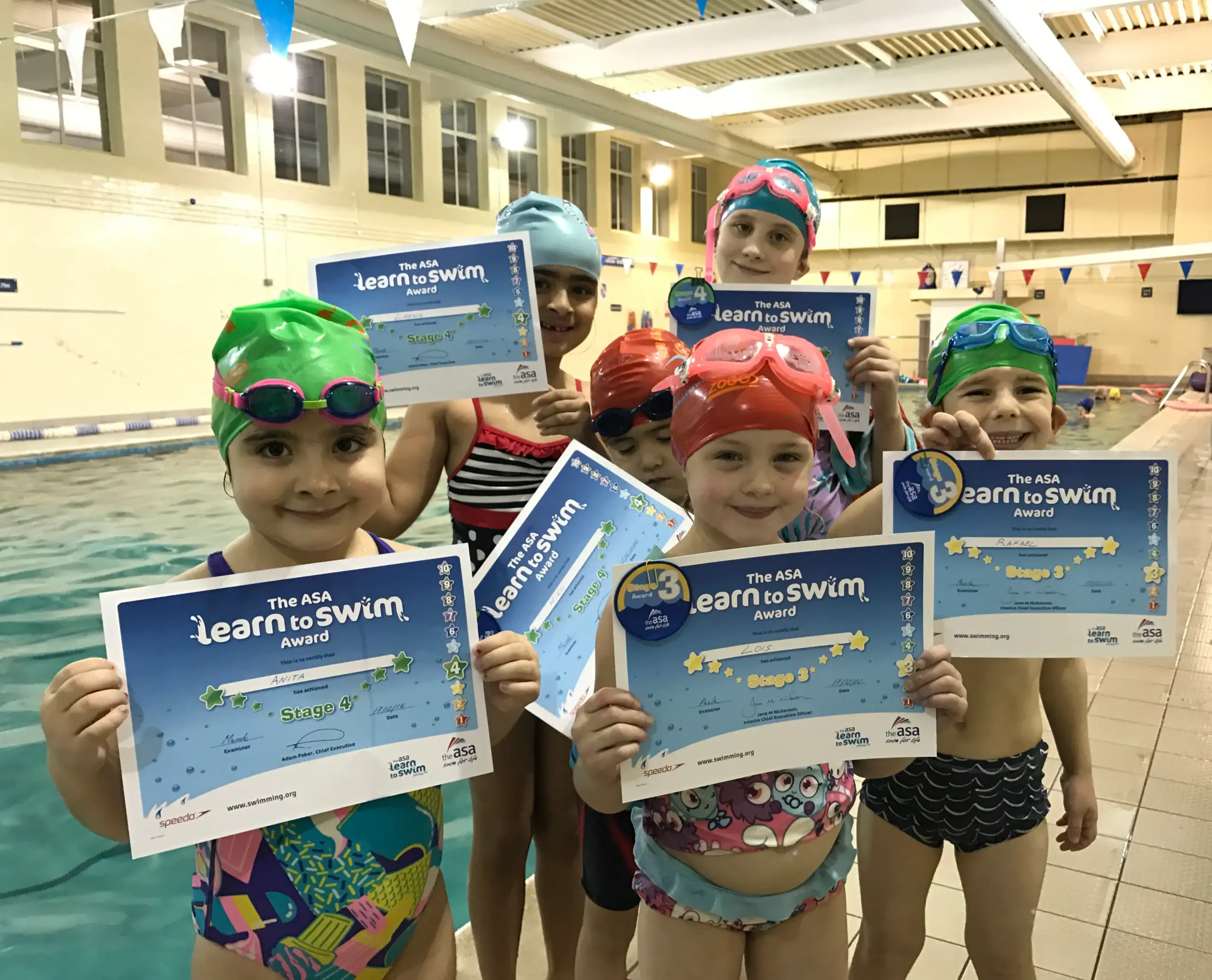 A group of happy children at Blue Wave Swim School holding their Swim England Learn to Swim Stage 3 and Stage 4 badges and certificates by the pool.