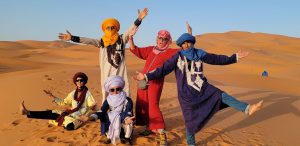 4 happy young men dressed in colorful robes and turbans stretch hands in the air while blogger UNSTOPPABLE Stacey reaches to the sky amongst them in the Sahara Desert