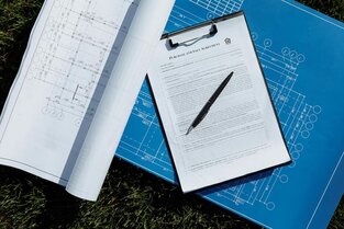 A purchase and sale agreement document on a clipboard with a pen rests atop architectural blueprints and rolled construction drawings, laid out on grass, symbolising the formal documentation required in contract novation processes.