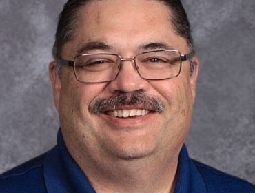 Headshot of man with dark mustache and glasses, wearing a blue polo shirt. He is smiling at the camera.