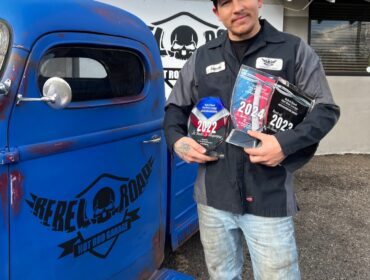 Man in WyoTech hat and shirt, holding three Business Hall of Fame awards. He is standing next to a royal blue truck that has Rebel Road Hot Rod Garage on side of the door.