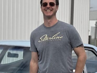 Man in sunglasses and grey t-shirt, standing next to a light blue car. Smiling at camera.