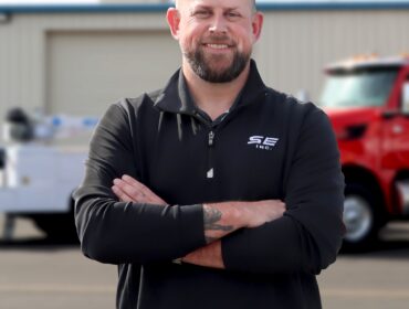 Man with brown goatee and a black pullover, smiling at the camera with arms crossed.