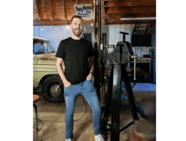 Man in brown beard, black t-shirt, and blue jeans, smiling at the camera. He is standing in a garage with a green, rusty truck behind him. To his left is a black vice. Behind everything is a work bench against the wall of the garage.