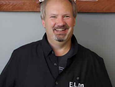 Man with greying goatee and black button up shirt, smiling at camera.