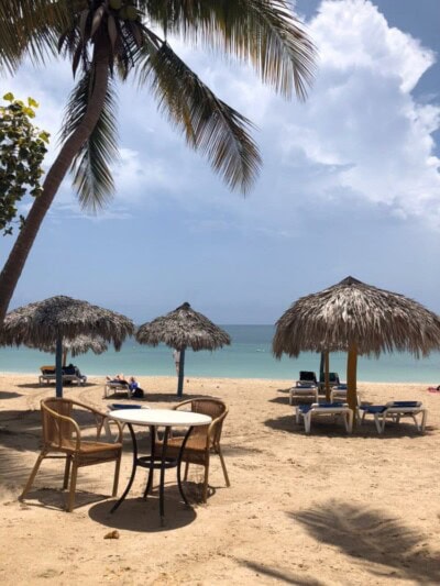 Sto i stolice ispod slamnatih suncobrana na plaži uz palmu i more