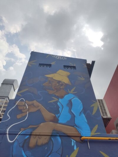 Mural na visokoj zgradi prikazuje starijeg muškarca u žutom šeširu i plavoj košulji