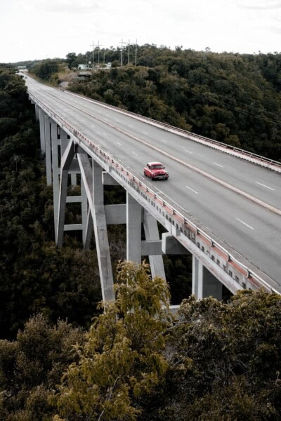 Crveni automobil prelazi preko visokog betonskog mosta iznad šume