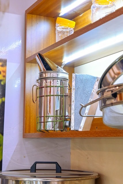 Metal beekeeping smoker displayed on a wooden shelf beside jars and honey-processing equipment.