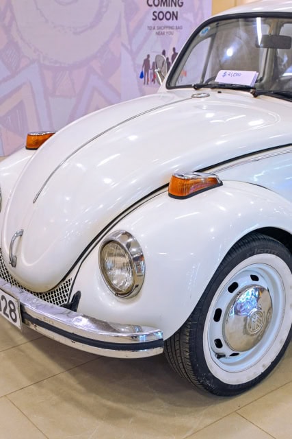 White vintage Volkswagen Beetle displayed indoors with a Coming Soon sign in the background.