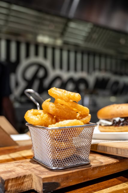 Golden fried onion rings served in a small metal basket with a burger in the background.