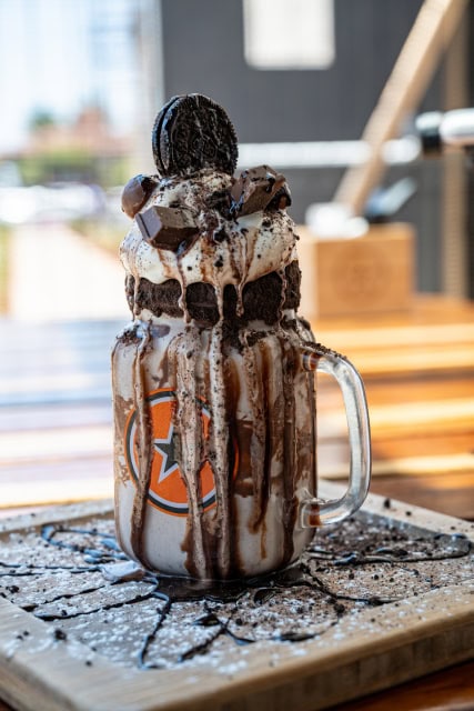 Tall chocolate milkshake in a handled glass mug topped with whipped cream, cookie crumbs and chocolate pieces.