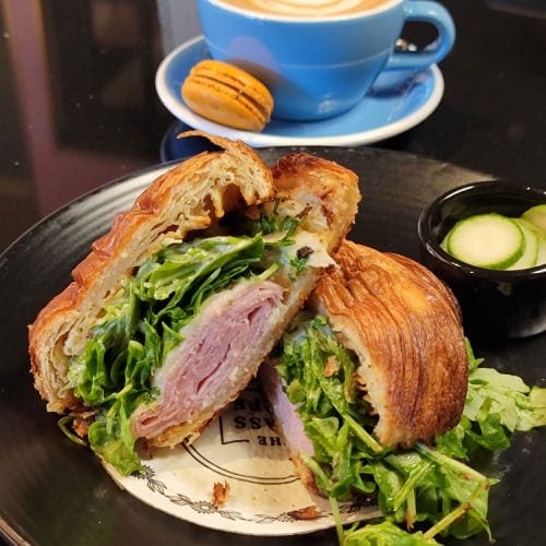 croissant sandwich cut in half filled with green arugula and ham on black table with blue coffee cup in background