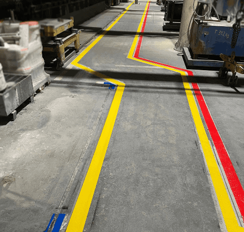 Fluchtwegmarkierung in RAL 1023 Verkehrsgelb und RAL 3020 Verkehrsrot. Der Fußweg wurde durch Kugelstrahlen vorbereitet, um eine bessere Haftung der Farbe zu gewährleisten. Die gelbe Markierung sorgt für sichere Gehwege für Mitarbeiter und Besucher, während die rote Markierung Gefahrbereiche abgrenzt und zur Verringerung von Unfällen beiträgt