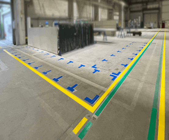 Blaue Markierungswinkel definieren Stellflächen an Maschinen in Halle mit gelb-grünem Linienraster.