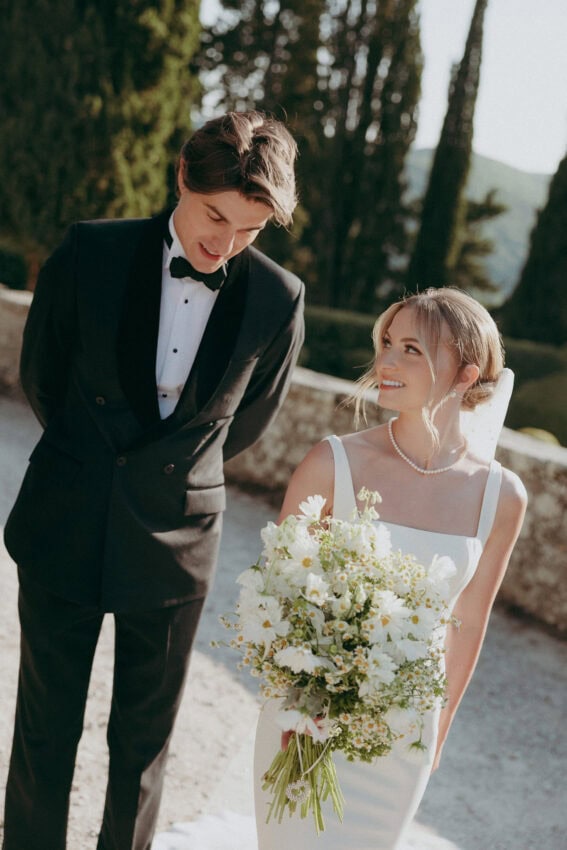 A bride in a white dress holding a bouquet of flowers smiles at her groom in a black tuxedo. They stand outdoors amid tall green trees and sunlight, with the picturesque Villa del Balbianello as their romantic backdrop.