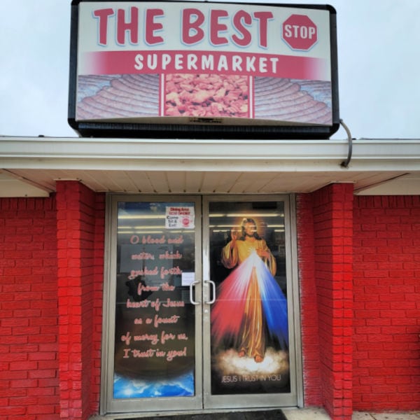 entry to smalltown market with two glass doors painted with Catholic images and the words 'Jesus i trust in you' with a sign on top of the roof reading 'The Best Stop Supermarket'