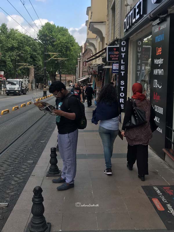 vacaywork ashrith standing next to tram line readin map and two turkish woman wearing denim, t-shirt and one of them in hijab passing by him