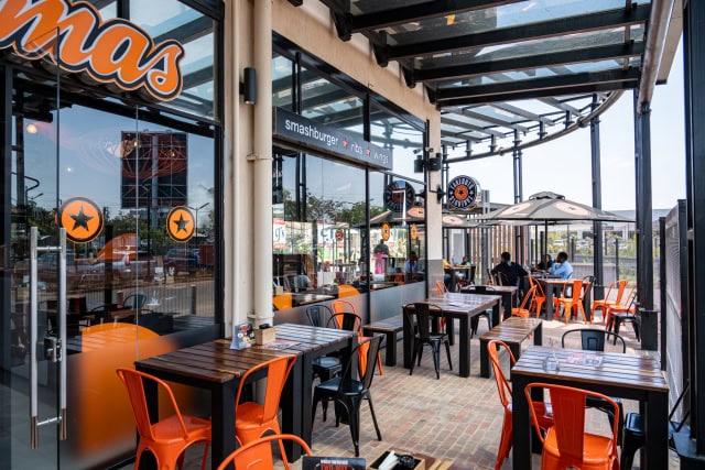 Wide covered dining terrace with rows of tables, orange chairs, glass storefronts, and outdoor umbrellas.