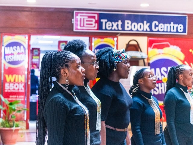 Women in black outfits sing in formation inside a shopping mall