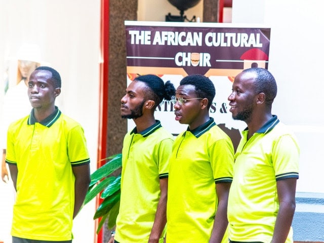 Four men in matching bright yellow polo shirts standing beneath a sign reading The African Cultural Choir