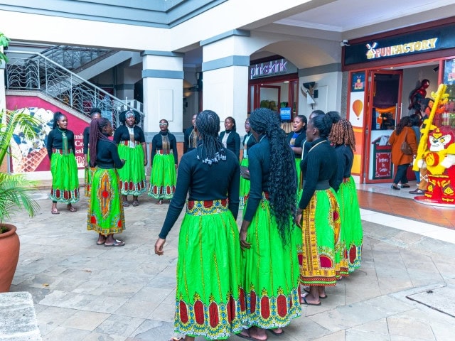 Women wearing bright green patterned skirts gather for a performance outside shops