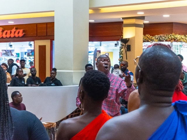 Man in a red plaid shirt sings or speaks to a crowd in a shopping mall