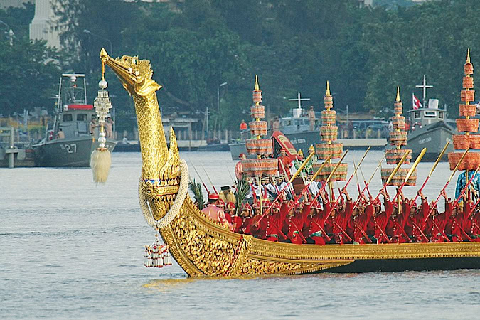 Gold lacquered wooden long boat with tall bow in the shape of a swan is paddled by dozens of red-clad oarsmen.