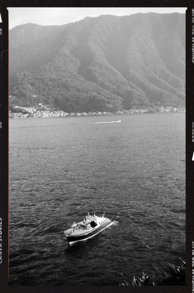 A black-and-white photo of a motorboat on a lake, with tree-covered mountains and a small town in the background. The scene is calm, with gentle waves on the water and a distant boat visible near the shoreline.