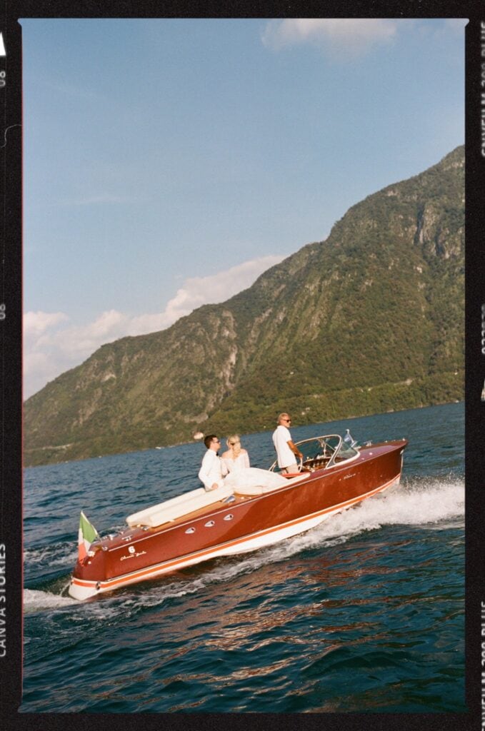 Four people ride in a vintage wooden speedboat on a lake, surrounded by mountains and blue sky. The water is calm and an Italian flag is visible on the boats stern. The group appears relaxed and is enjoying the scenery.