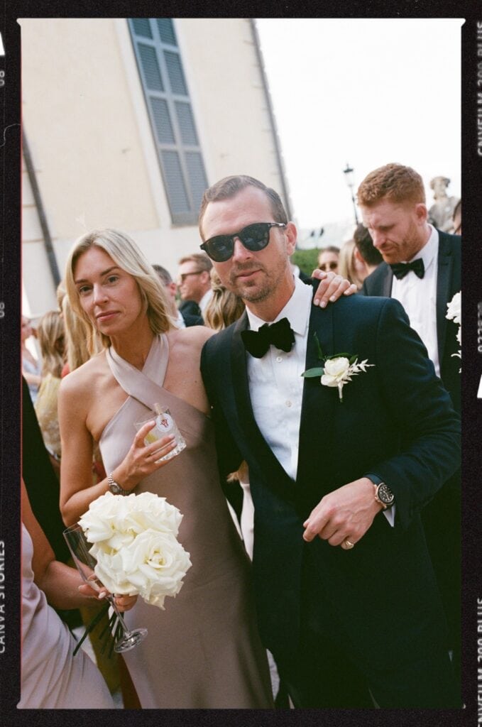 A well-dressed man in a tuxedo and sunglasses poses with a woman in a light beige, one-shoulder dress holding a bouquet of white roses at a formal outdoor event, surrounded by other guests.