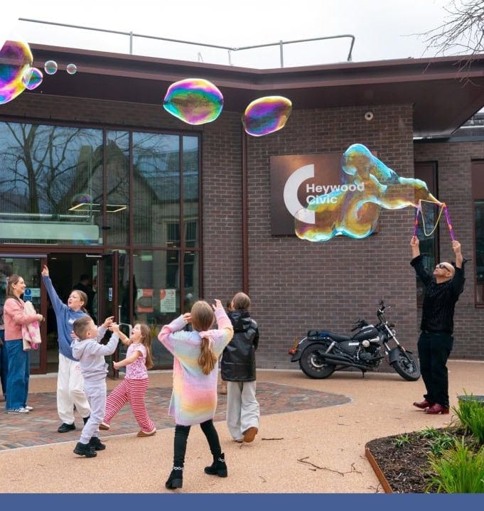 an external photo of heywood civic centre entrance with a performer making giant bubbles with children playing trying to catch them to the rear of the image there is a parked black motorbike