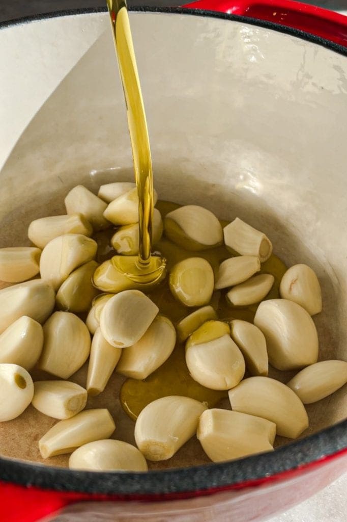 This Garlic Confit recipe is going to become your new pantry staple because it's so easy and confited garlic is so spectacular and versatile. You just need a few heads of garlic, your favourite spices and herbs, lots of olive oil and 30 minutes. The result is a scrumptious sort of sweet spreadable garlic that you can use in a thousand ways. As a bonus, you get garlic-infused oil! In this picture you can see the raw garlic in a cocotte being covered with olive oil. | Paula's Apron Recipe