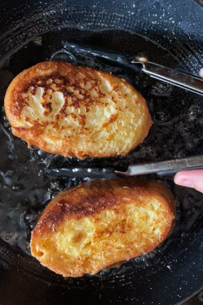 These Traditional Torrijas or Spanish Style French Toasts are homemade and so easy to make. They are creamy on the inside and have a cinnamon sugar coating. Plus, they are the perfect excuse to make the most of stale bread. If you love French toasts, you will love this delicious Spanish Easter dessert. In this shot you can see 2 torrijas being fried and already golden on the side facing us. | Paula's Apron Recipe