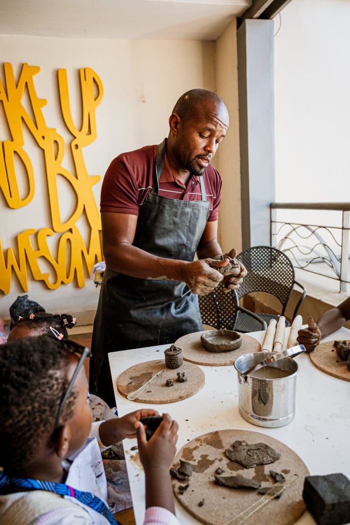 An instructor demonstrates clay work to children gathered around a workshop table.