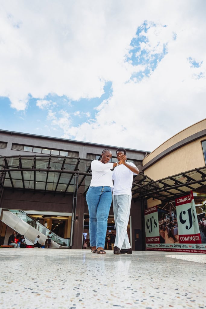 Two dancers walk together through a mall courtyard near a glass entrance