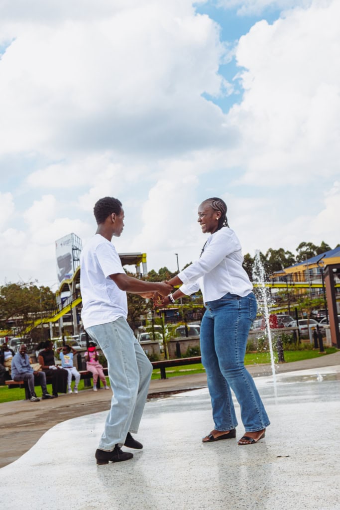 Two dancers hold hands while performing on the wet courtyard surface beside the mall fountain.