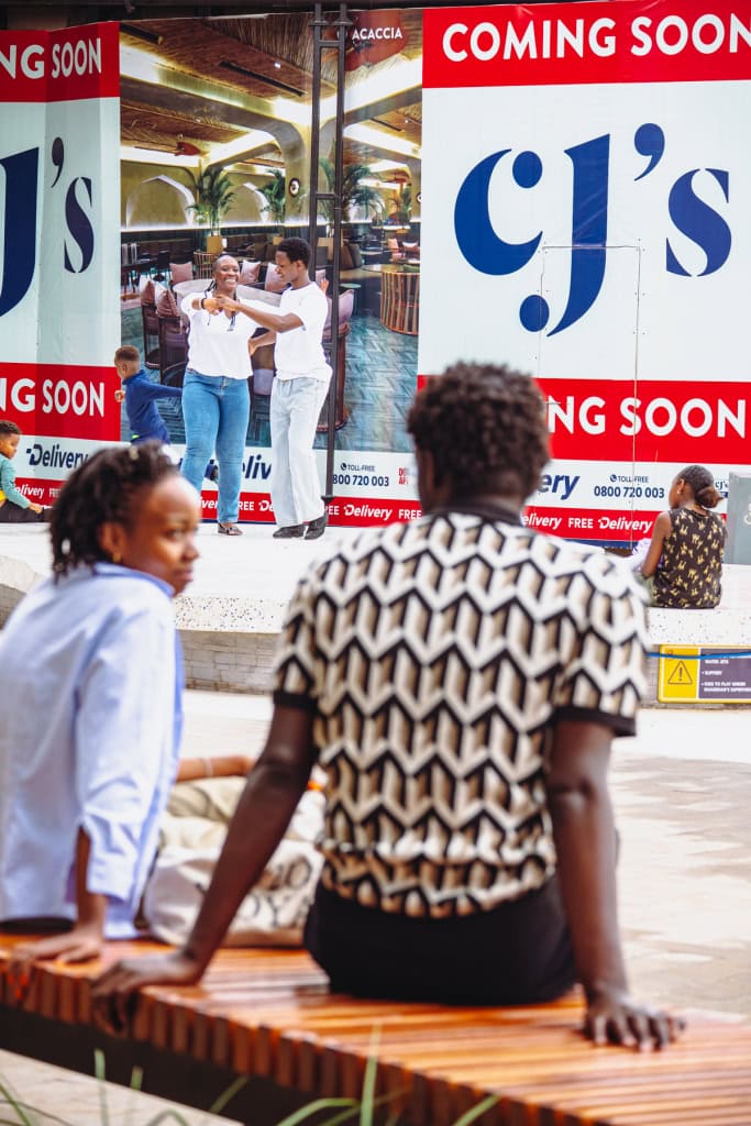 Two dancers are visible behind seated shoppers and large restaurant signage at the mall