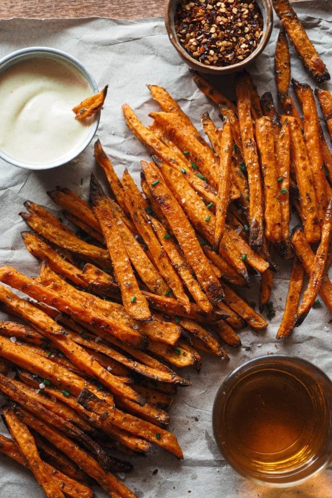 Crispy Baked Sweet Potato Fries with Spicy Mayo, the perfect fall appetizer, side dish, game-day snack, it all works. The cool thing is that they’re healthier than if fried, super easy to make and are actually crispy! There's a small bowl with red pepper flakes at the top, a metallic bowl with the spicy mayo and a dipped sweet potato on the left and a glass with beer at the bottom. In the centre and diagonally the sweet potatoes are laying.