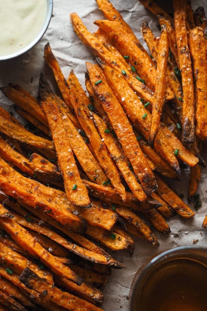 Crispy Baked Sweet Potato Fries with Spicy Mayo, the perfect fall appetizer, side dish, game-day snack, it all works. The cool thing is that they’re healthier than if fried, super easy to make and are actually crispy! The sweet potatoes can be seen zoomed in with the flaky sea salt and chopped chives. They are laying between a bowl with the dip and a glass of beer.